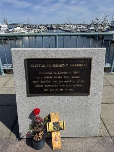 Seattle Fishermen's Memorial plaque honoring lives lost at sea, flowers at its base beside a marina full of boats.